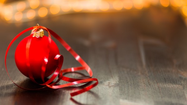 Zu sehen ist eine rote Weihnachtsbaumkugel mit einer goldenen Krone an einem roten Band. Die Kugel liegt auf einem dunklen Parkettboden und im Hintergrund sind verschwommene Lichter zu erkennen.